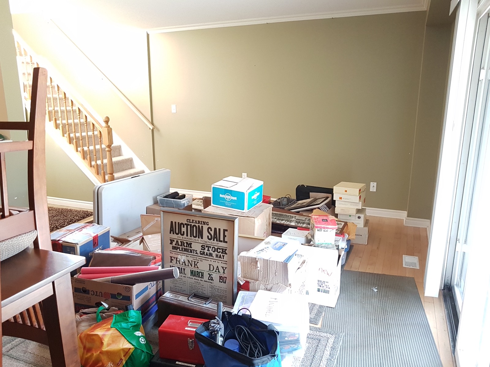 A room mid-move: boxes, bags and furniture stacked by the stairs, light coming in from the patio door.